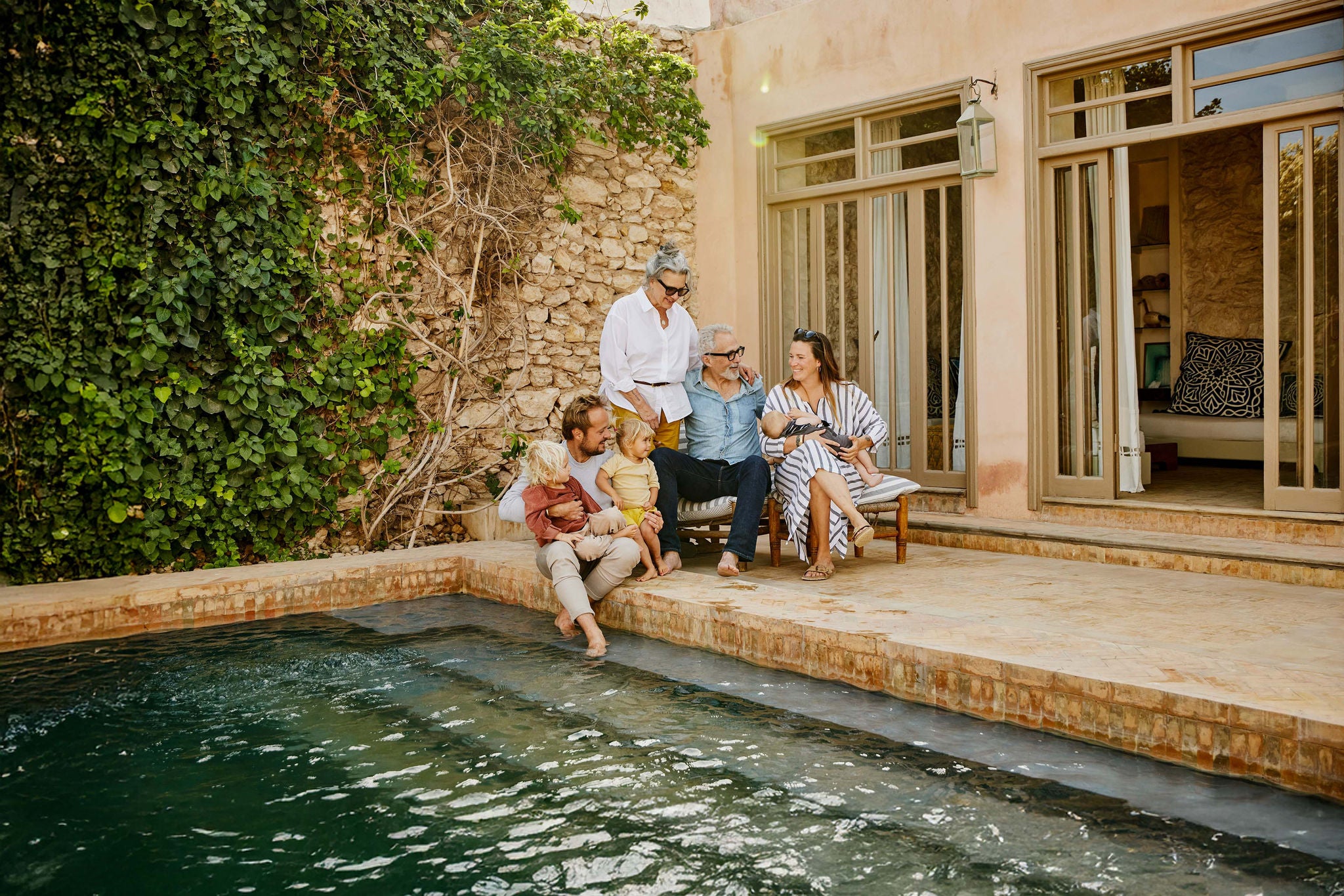 Wide shot of smiling grandparents relaxing with children and grandchildren beside pool at tropical villa during multigenerational family vacation