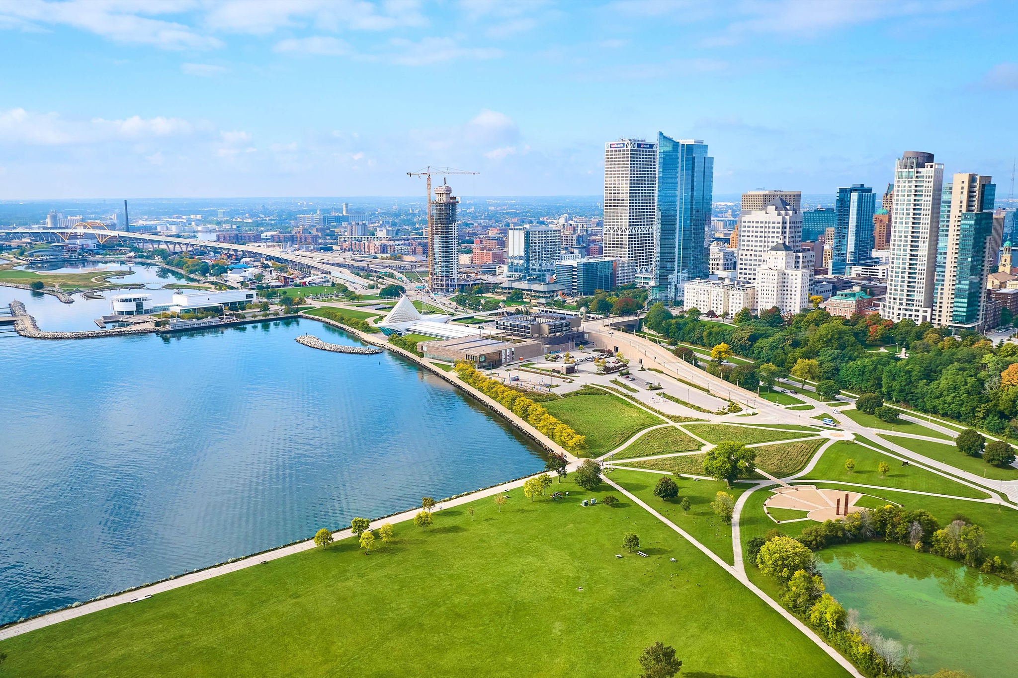 Drone view of Milwaukee's vibrant cityscape, showcasing the contrast between lush parklands and modern high-rise buildings, with iconic Quadracci Pavilion and Lake Michigan in view.