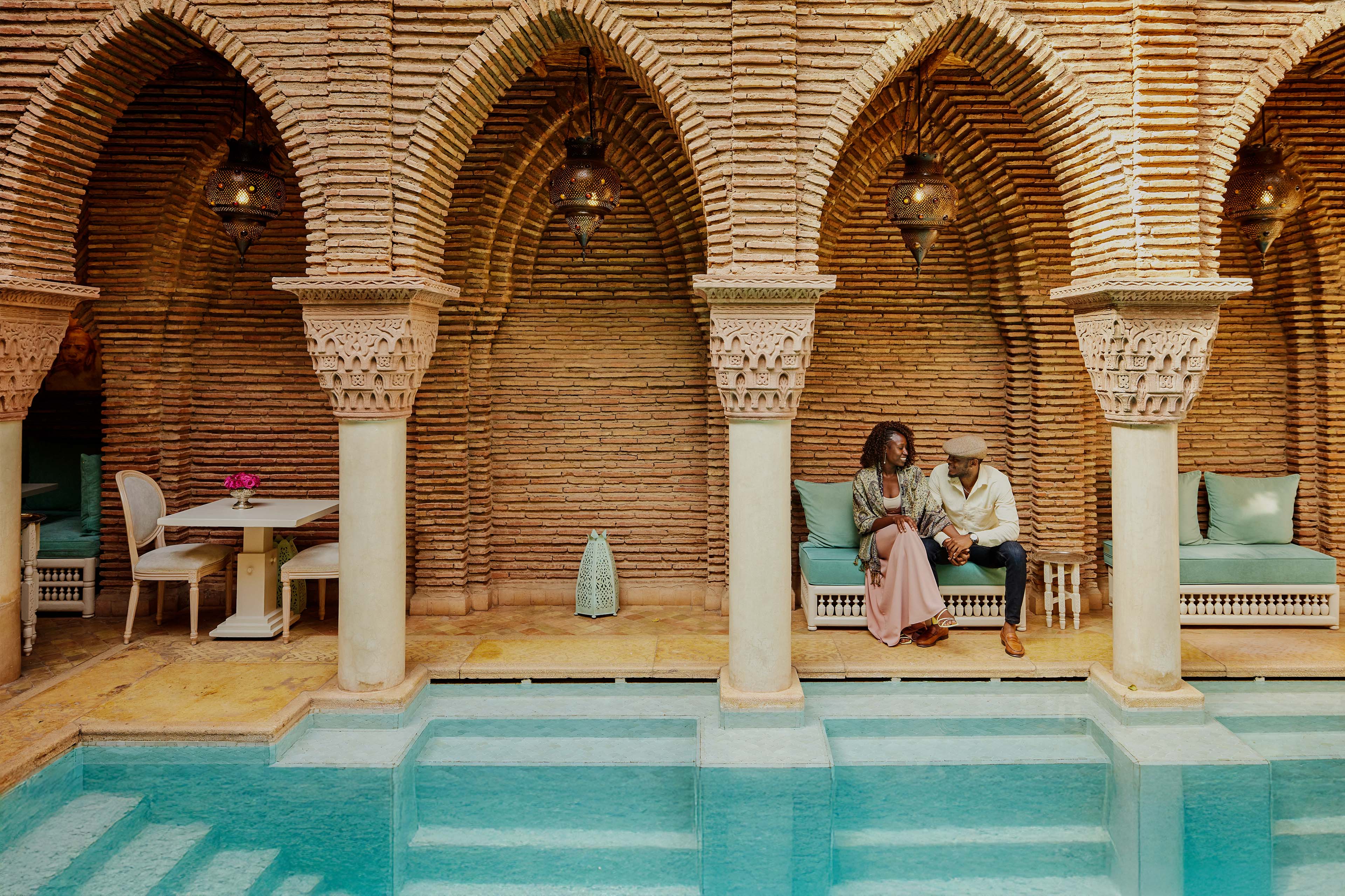 Wide shot of smiling couple holding hands while sitting by pool in courtyard of luxury hotel while on vacation